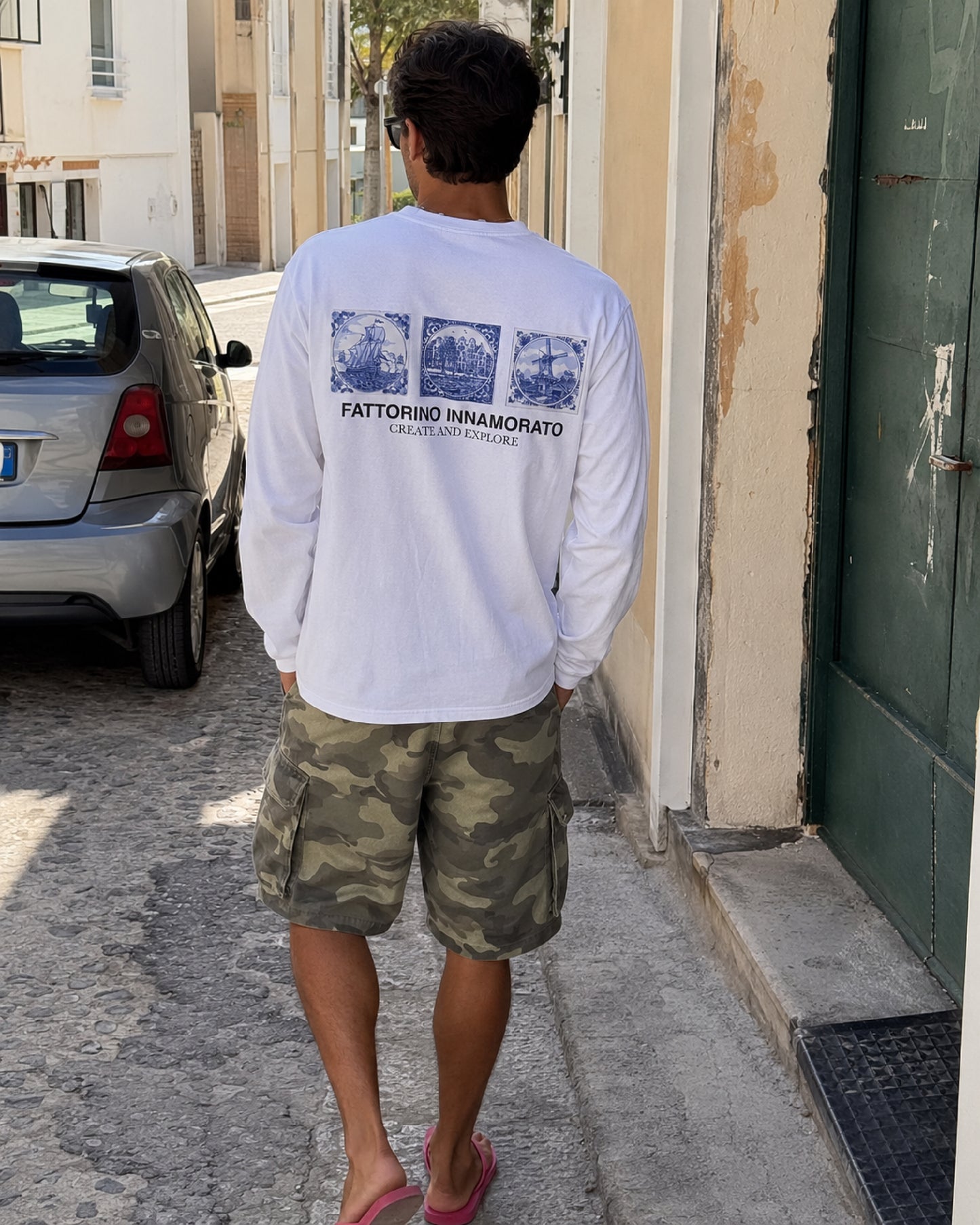 A man viewed from behind walking down a sunlit street, wearing a white long-sleeve t-shirt with a blue graphic print on the back and green camouflage cargo shorts. Dutch oversized Longsleeve Fattorino Innamorato Create and explore