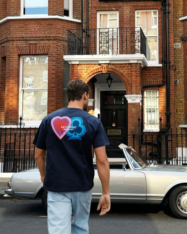 Person wearing a dark blue t-shirt with colorful designs walking past a classic car and brick building. Fattorino Innamorato. Play navy

