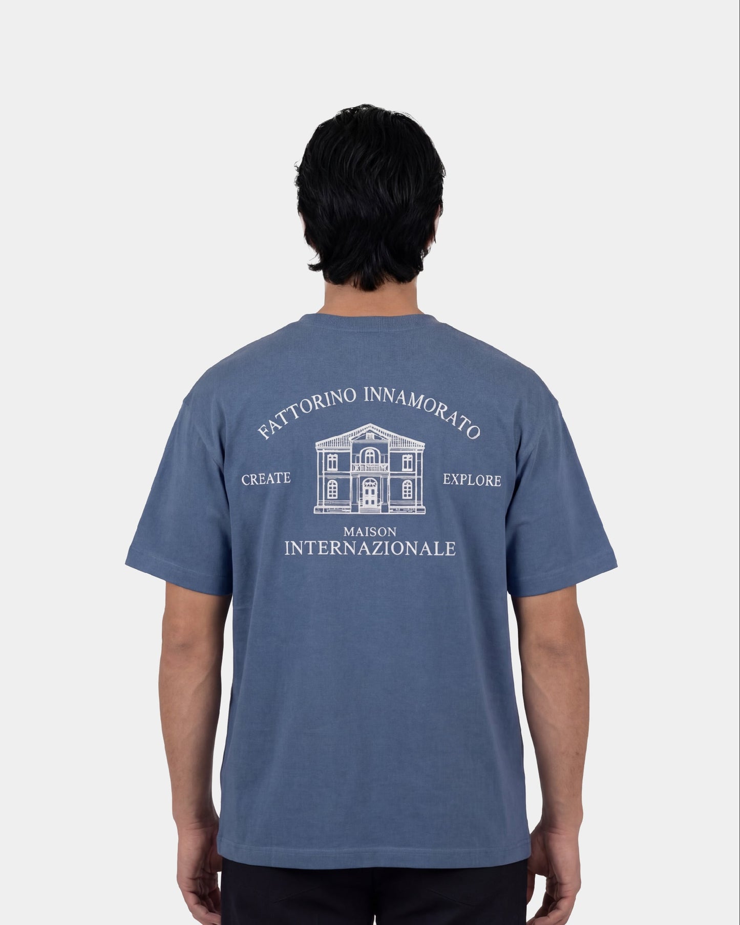 Blue t-shirt with white text and building graphic on a light gray background. Fattorino innamorato maison blue