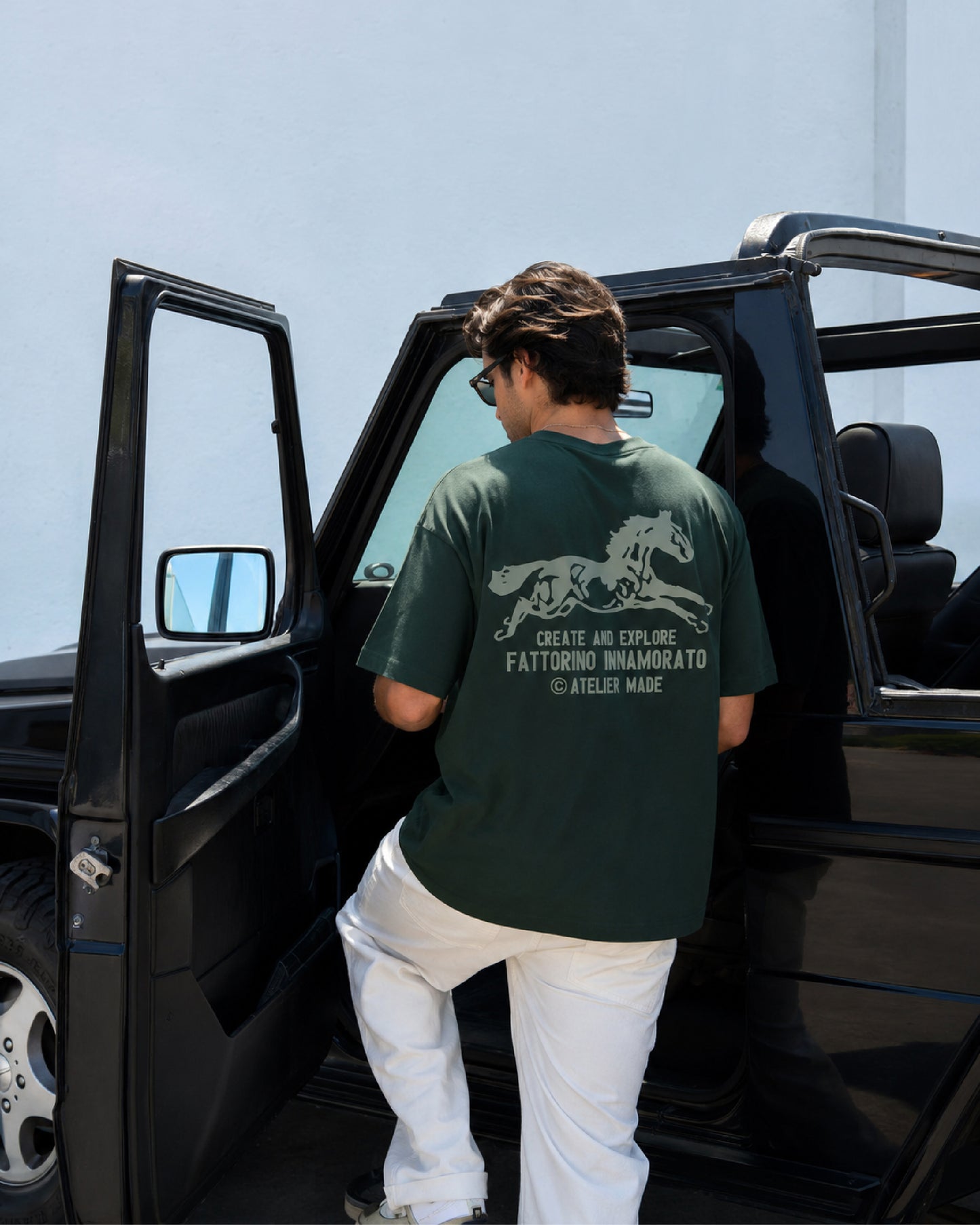 A man wearing a dark green t-shirt with a white graphic print on the back, standing in the doorway of a black SUV. Fattorino Innamorato Cavallo Green