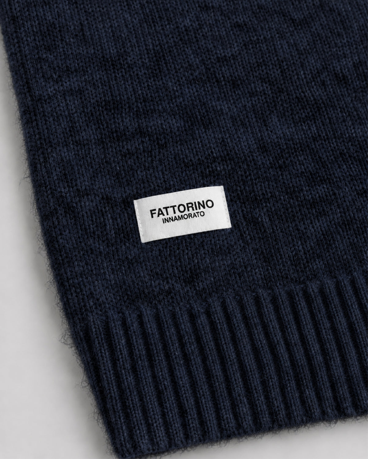 Close-up of a dark navy knit fabric with a white rectangular label reading FATTORINO INNAMORATO, showing the ribbed cuff edge against a light neutral background.