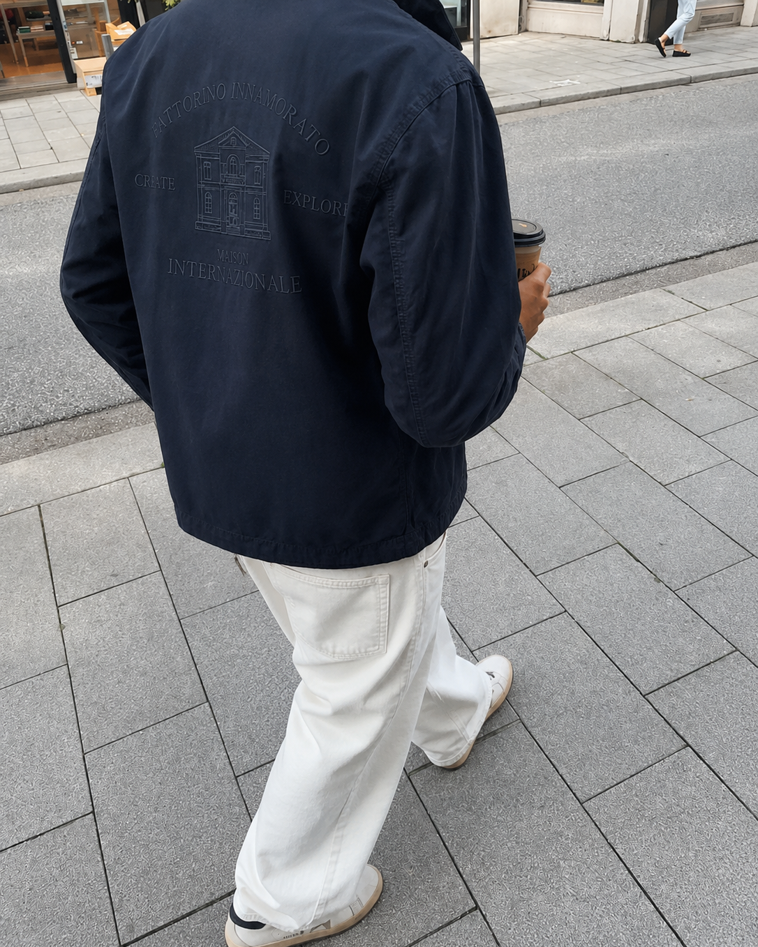 A person viewed from behind wearing a dark navy jacket with tonal back embroidery, white pants, and white sneakers while walking on a gray paved sidewalk. Navy Fattorino Innamorato maison workwear jacket.