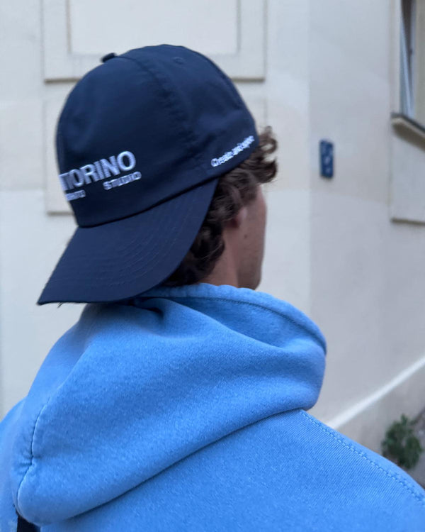 Person wearing a blue cap with 'Fattorino Innamorato Studio' branding, standing against a light-colored wall. Navy cap